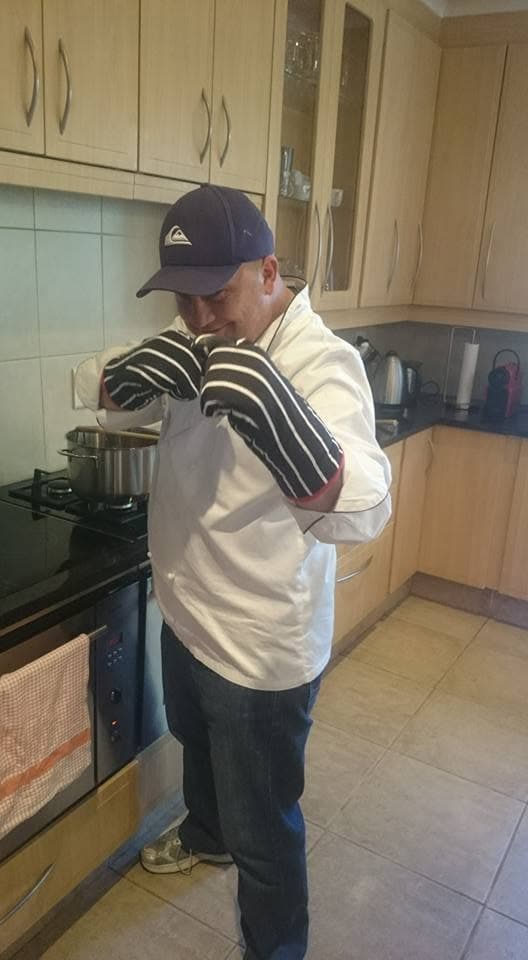 Paul in a home kitchen in his whites and a cap, oven gloves on, lifting the lid of a pot on the stove